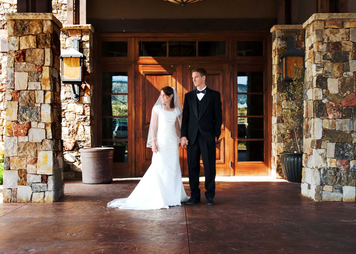 Bride and groom holding hands in front of tall wooden doors framed by stone pillars at The Ledges